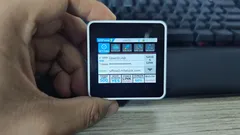 Hand holding a small touchscreen device displaying icons with a keyboard in the background